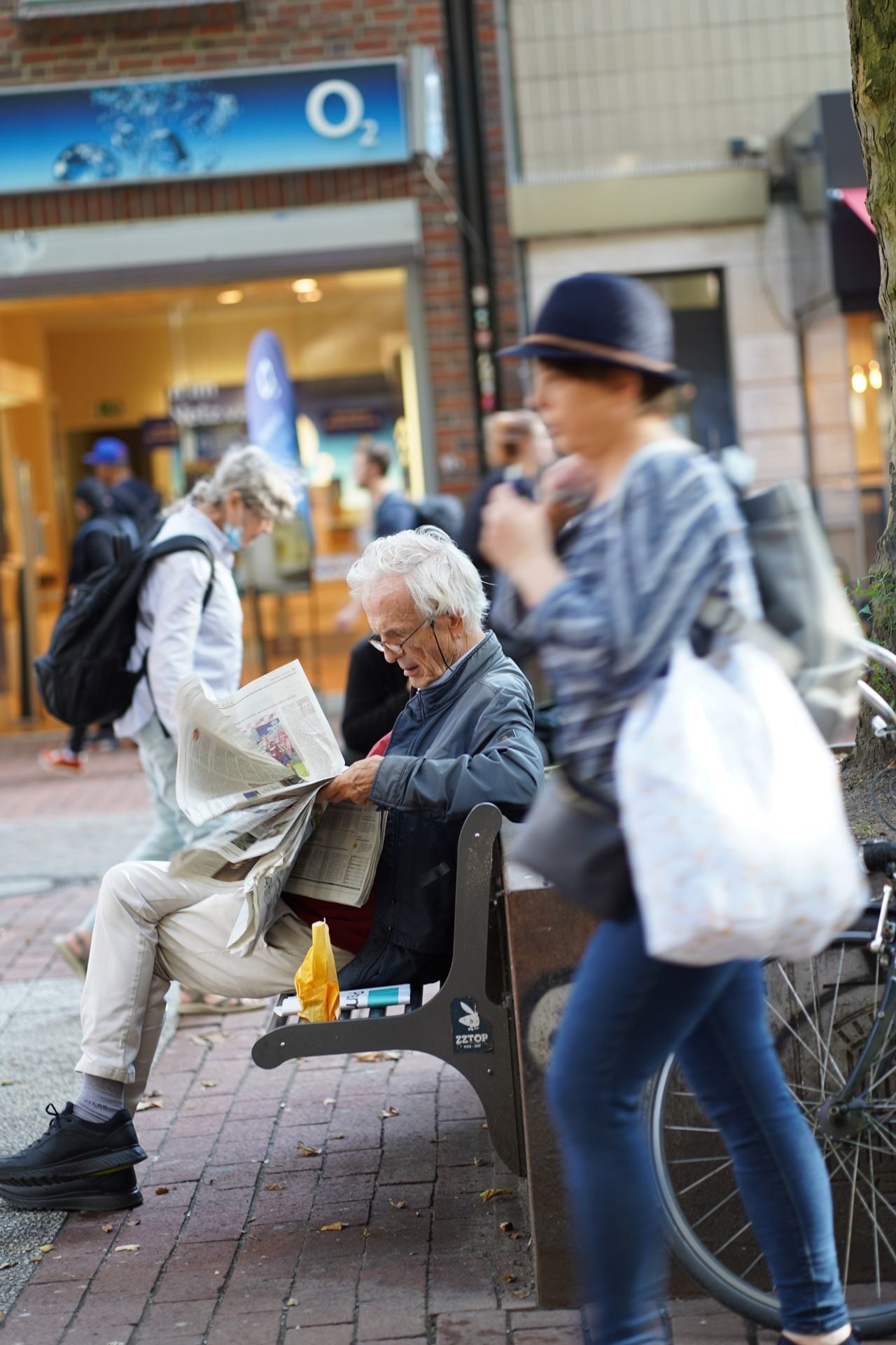 Senior man in eyeglasses reads a newspaper on a street bench with pedestrians and stores in the background.