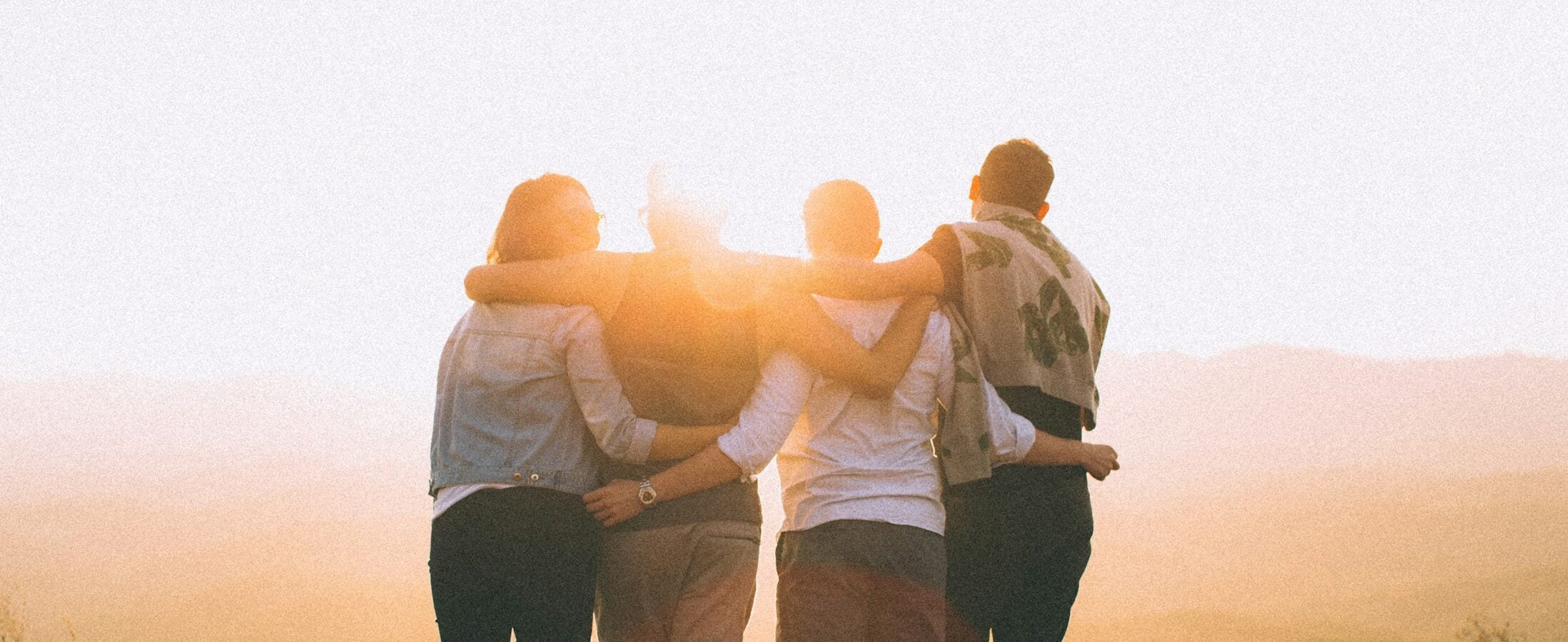 four person hands wrap around shoulders while looking at sunset